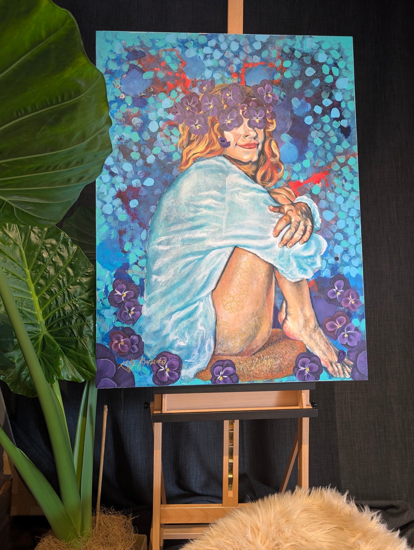 Full view of She Sat With Silence II by Kelly Batsiokis displayed on an easel beside a large elephant ear plant — original mixed media painting showing scale in a natural, creative studio setting.