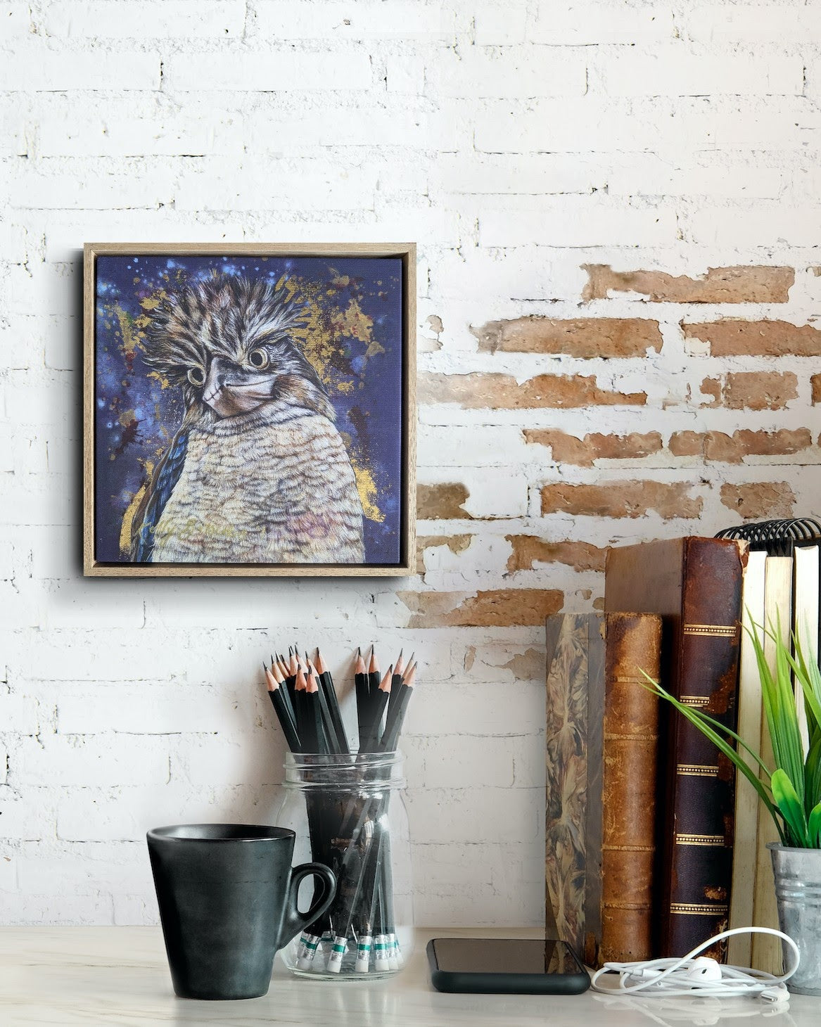 Framed fine art reproduction print of Bazza the Kookaburra by Kelly Batsiokis on a white brick wall with stationery items below for scale.