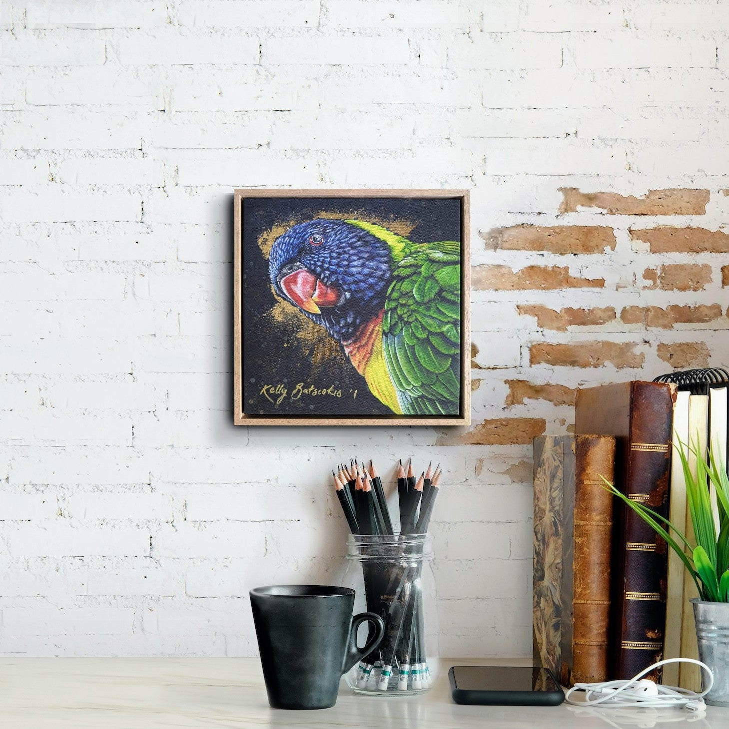 Framed fine art embellished print of Shazza the Rainbow Lorikeet on a white brick wall with stationery items below to show scale.