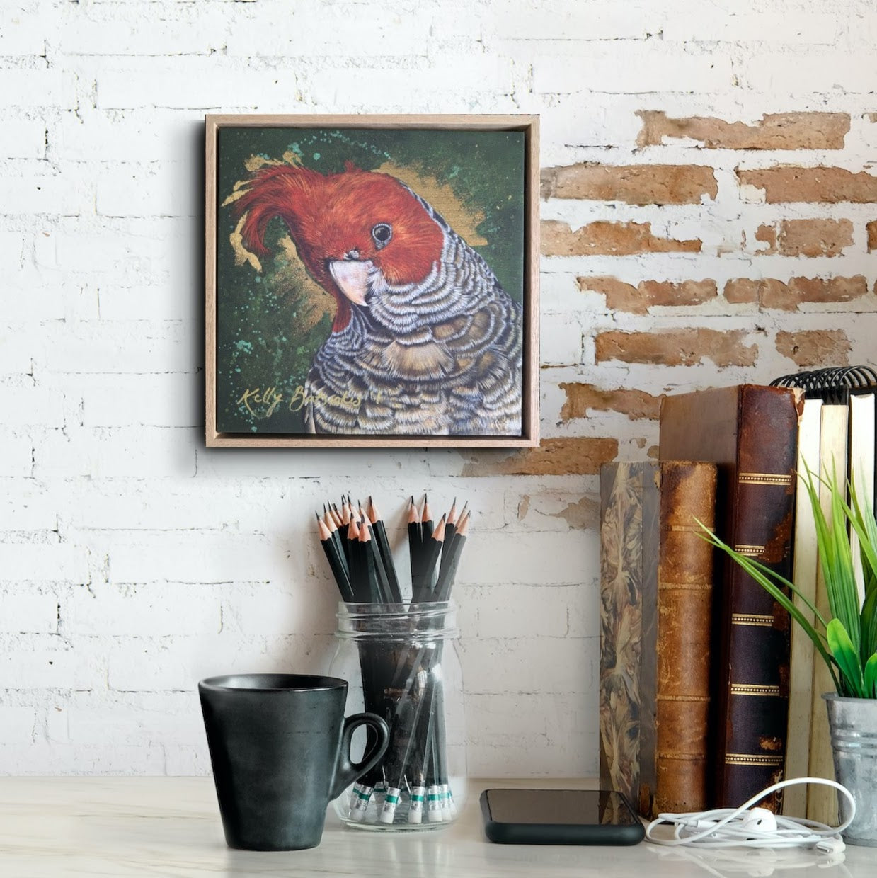 Framed, embellished fine art print of Elvis the Gang Gang Cockatoo by Kelly Batsiokis on a wall above a desk with stationery items for scale.