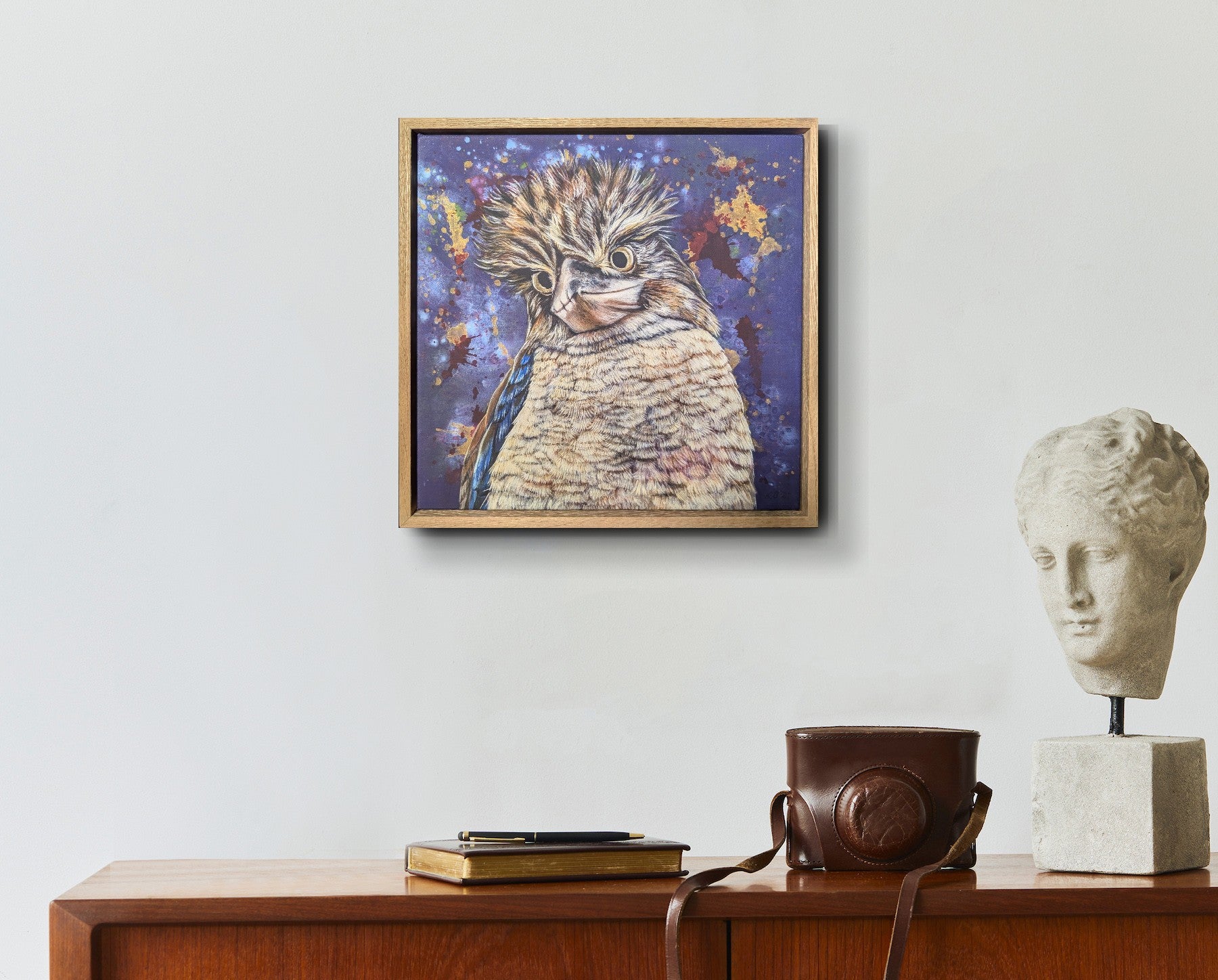 Framed fine art reproduction print of Bazza the Kookaburra by Kelly Batsiokis on a wall above a wooden cabinet with a camera and sculpture for scale.