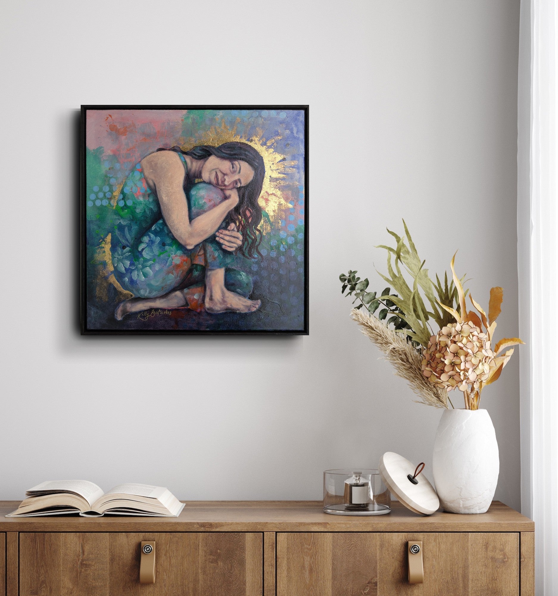 Framed artwork of a woman embracing a sun on a wall above a wooden cabinet with decor items.
