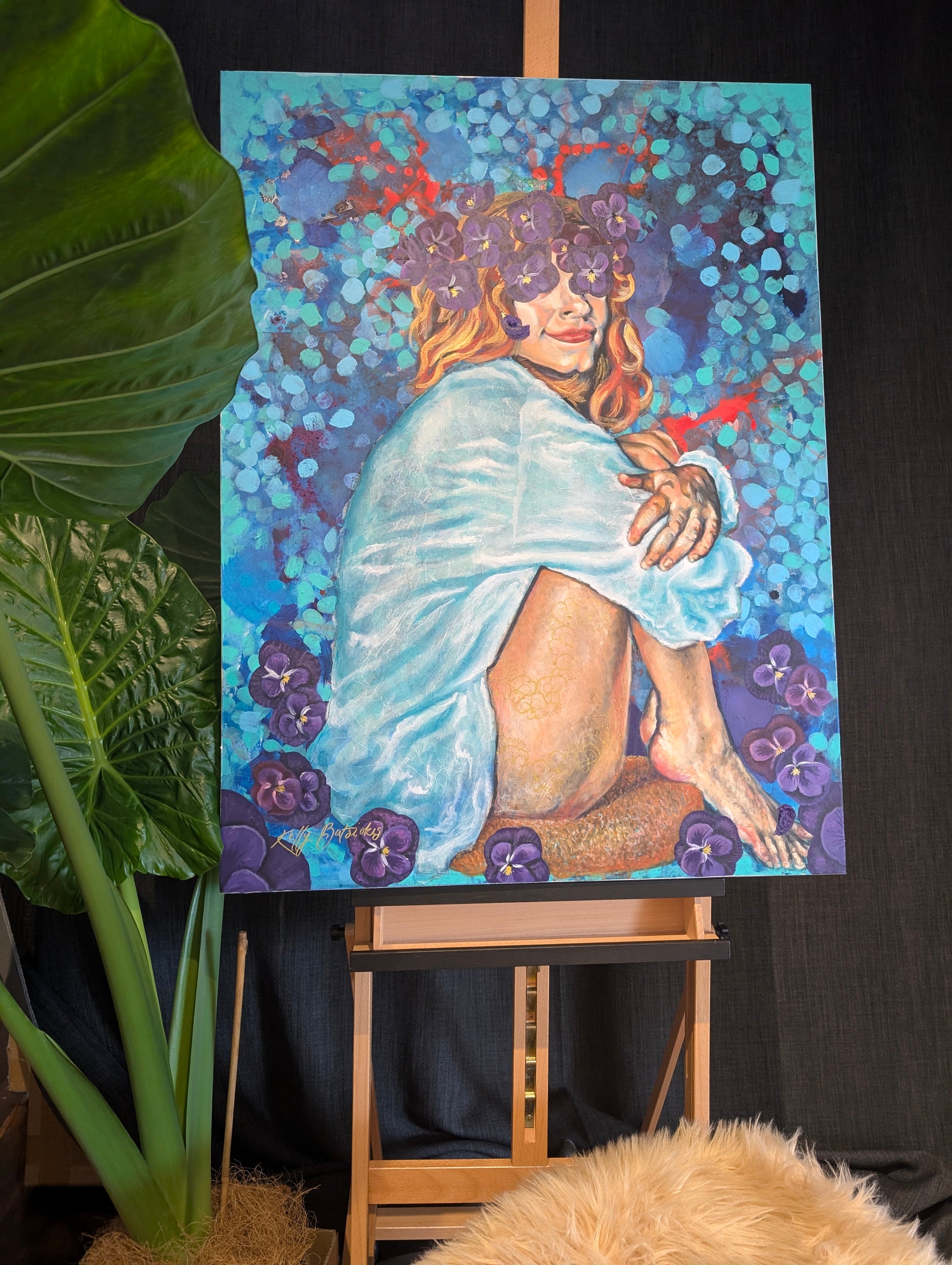 Full view of She Sat With Silence II by Kelly Batsiokis displayed on an easel beside a large elephant ear plant — original mixed media painting showing scale in a natural, creative studio setting.